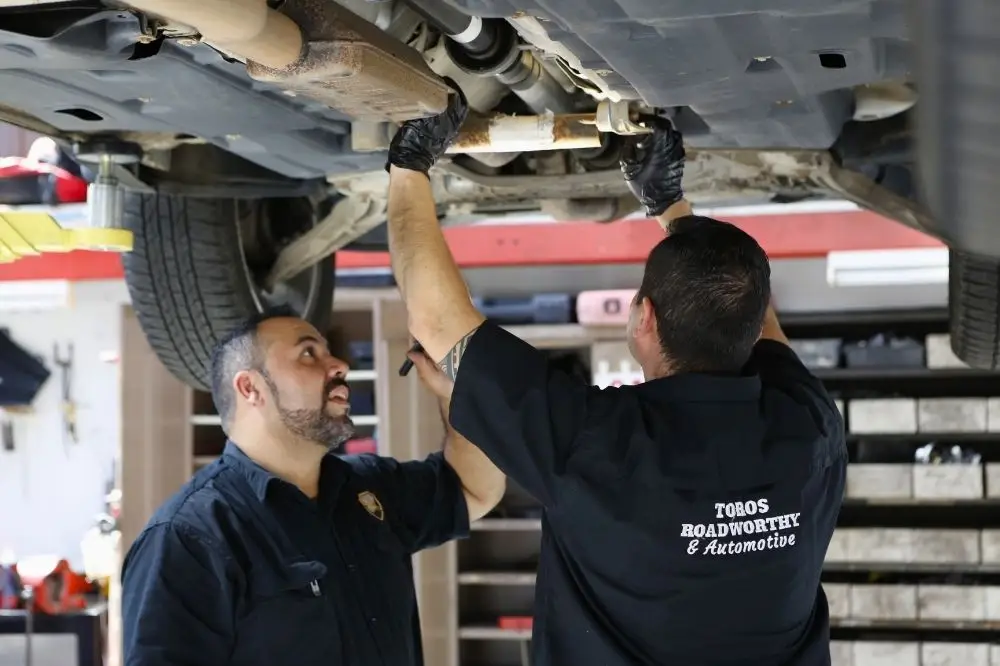 Mechanics performing roadworthy inspection under vehicle in workshop