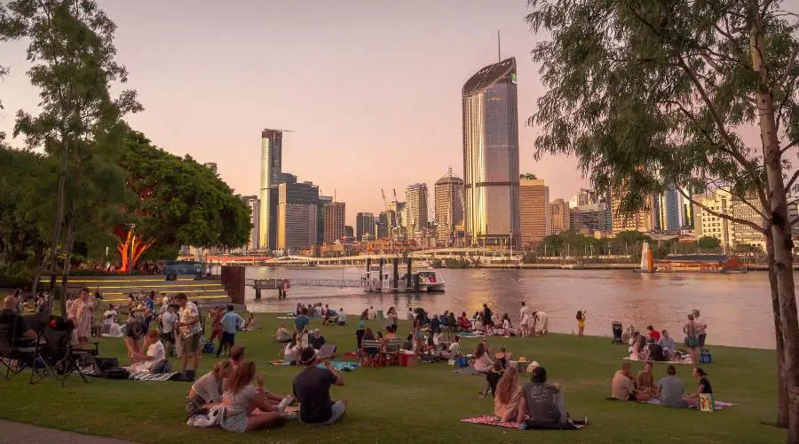 People relaxing in riverside park at sunset, city skyline.