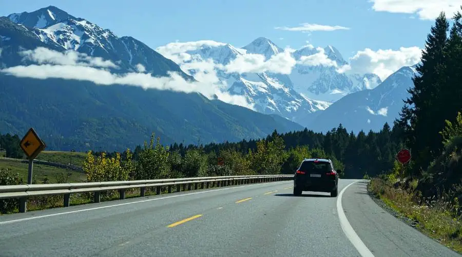 Car driving toward snow-capped mountains on sunny day.